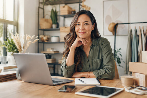 Smiling woman in a green shirt sits at a wooden desk with a laptop and notebook in a bright boutique workspace with a clothing rack nearby.