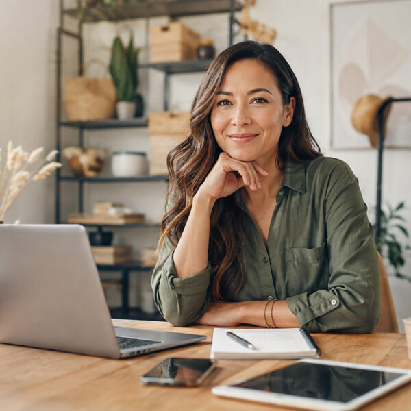 Smiling woman in a green shirt sits at a wooden desk with a laptop and notebook in a bright boutique workspace with a clothing rack nearby.